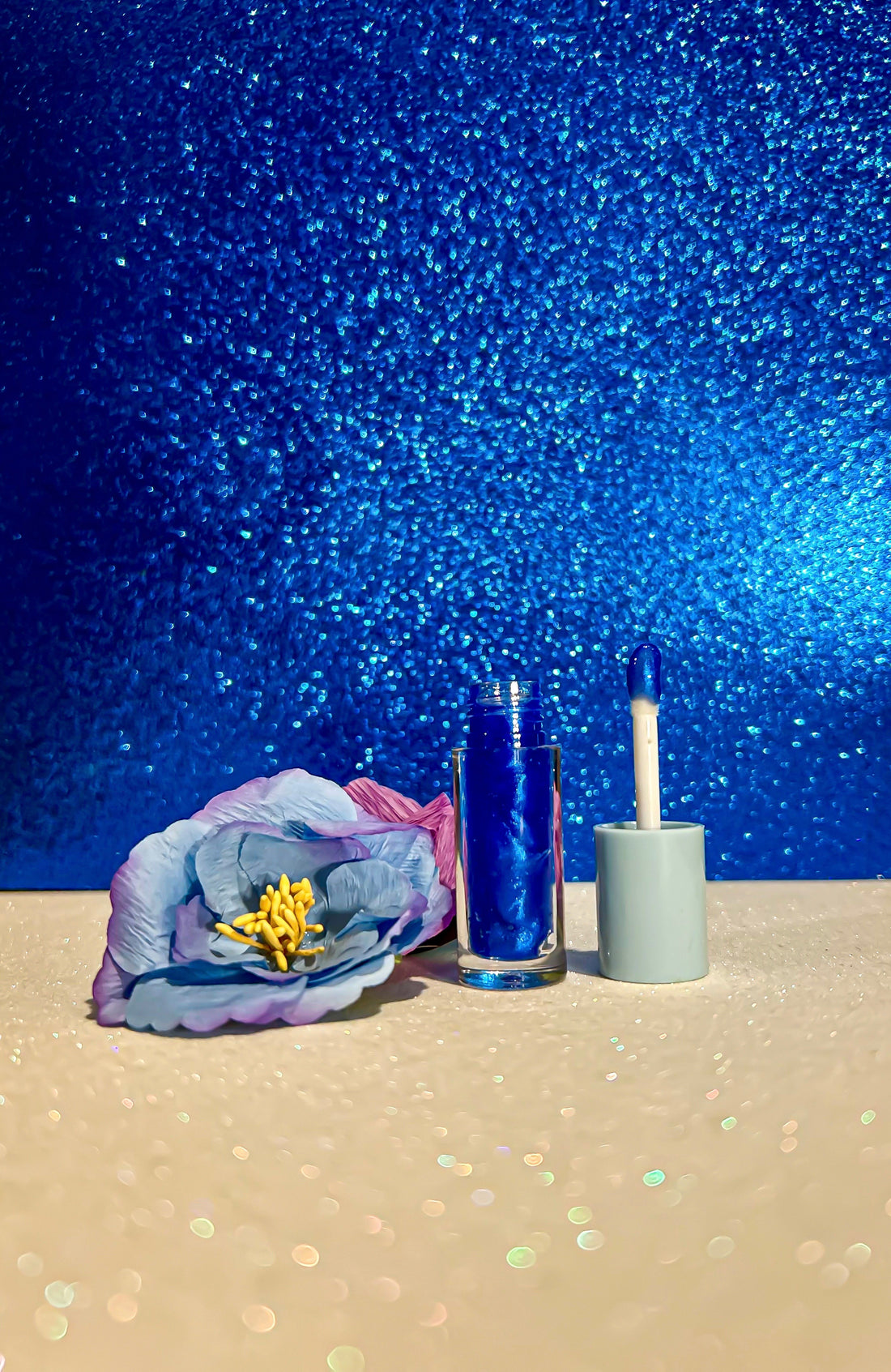 Coral Flavors Lip Gloss with applicator next to a blue and purple flower on a sparkly background.