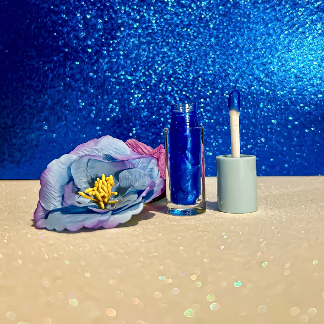 Coral Flavors Lip Gloss with applicator next to a blue and purple flower on a sparkly background.