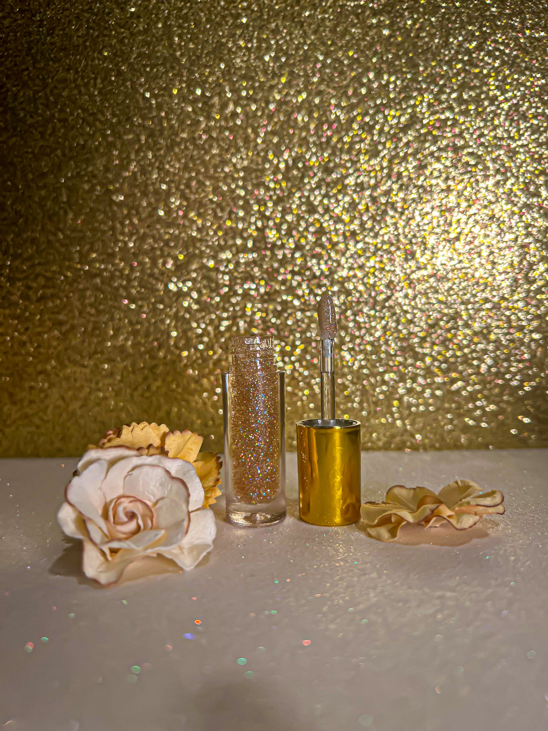 Luxe Flavors Lip Gloss in chic gold packaging beside decorative flowers on shimmering background.