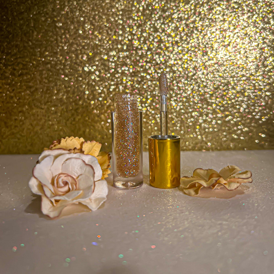 Luxe Flavors Lip Gloss in chic gold packaging beside decorative flowers on shimmering background.